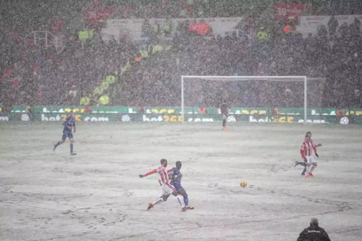 Jugadores de fútbol compiten en un campo cubierto de nieve durante un partido con una multitud de espectadores en las gradas.