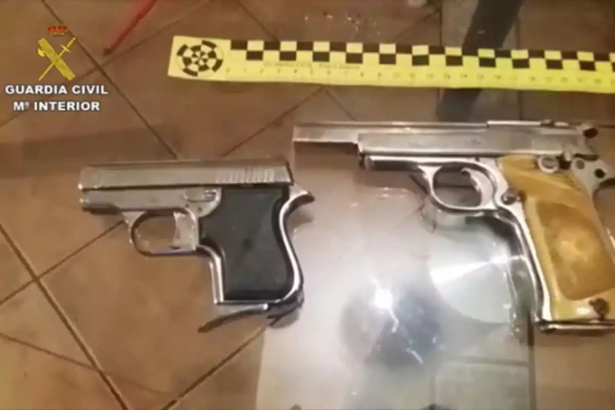 Dos pistolas de metal sobre una superficie de vidrio junto a una regla de medición amarilla y negra con el logotipo de la Guardia Civil en la esquina superior izquierda.