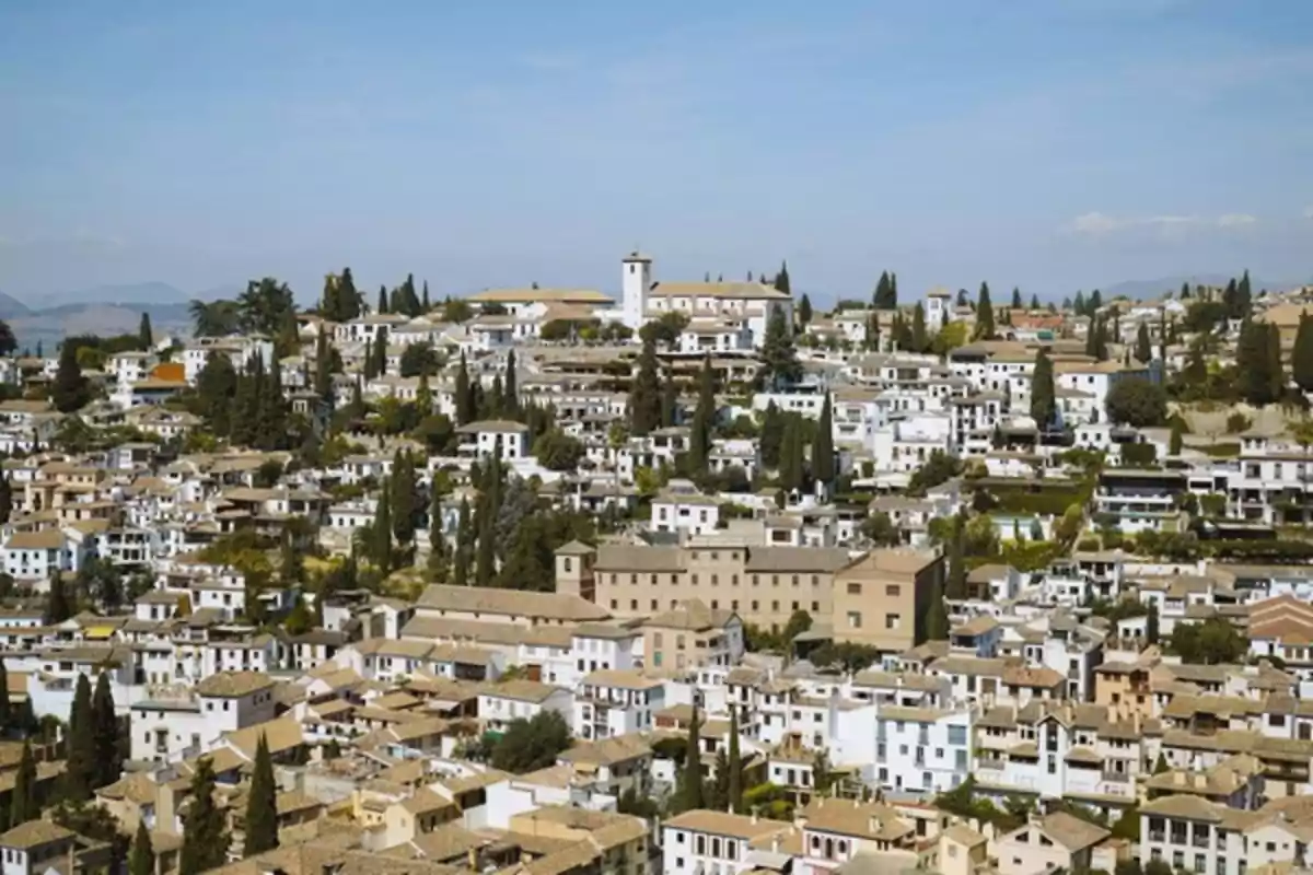 Vista panorámica de un barrio histórico con casas blancas y tejados de teja rodeadas de cipreses y vegetación bajo un cielo despejado.