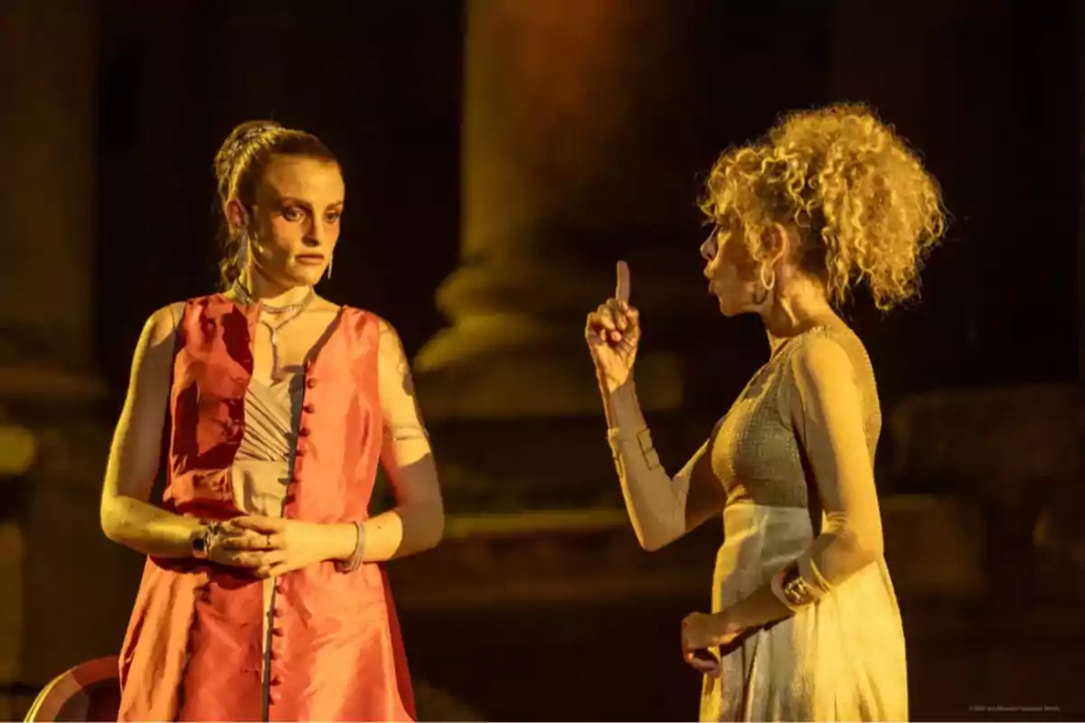 Dos mujeres en escena teatral con vestuario llamativo una de ellas gesticula con el dedo levantado mientras la otra la observa con expresión seria Dos mujeres en escena teatral con vestuario llamativo una de ellas gesticula con el dedo levantado mientras la otra la observa con expresión seria