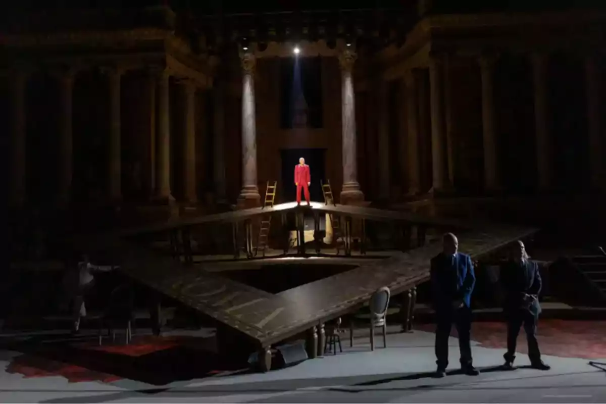 Escenario teatral oscuro con una persona vestida de rojo en el centro iluminada por un foco, rodeada de columnas y varias sillas, mientras dos personas de pie observan desde el primer plano Escenario teatral oscuro con una persona vestida de rojo en el centro iluminada por un foco, rodeada de columnas y varias sillas, mientras dos personas de pie observan desde el primer plano