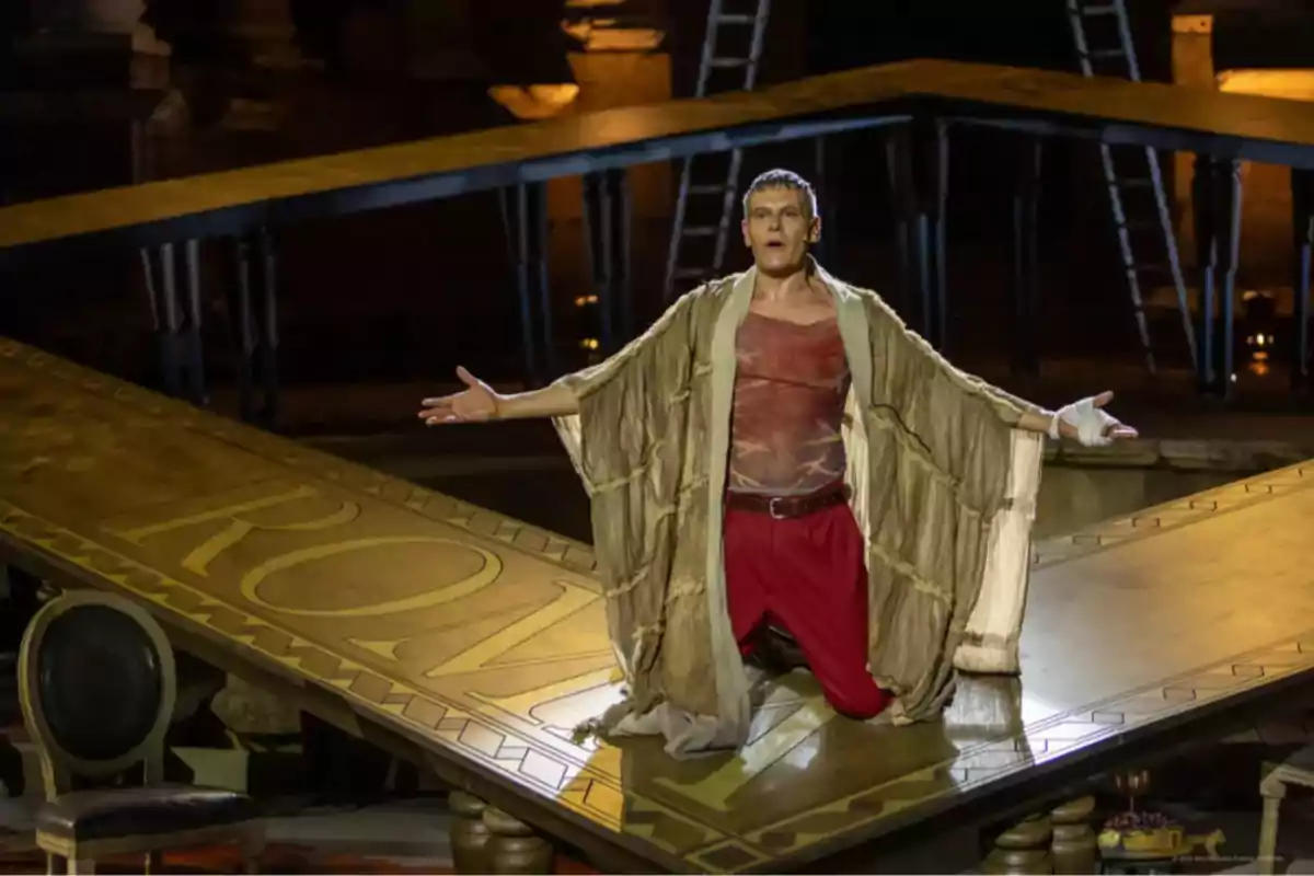 Actor arrodillado en el escenario con los brazos extendidos durante una obra teatral ambientada en la antigua Roma