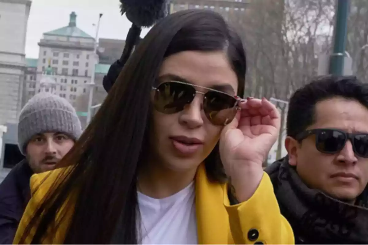 Mujer con gafas de sol y abrigo amarillo caminando por la calle acompañada de dos hombres.