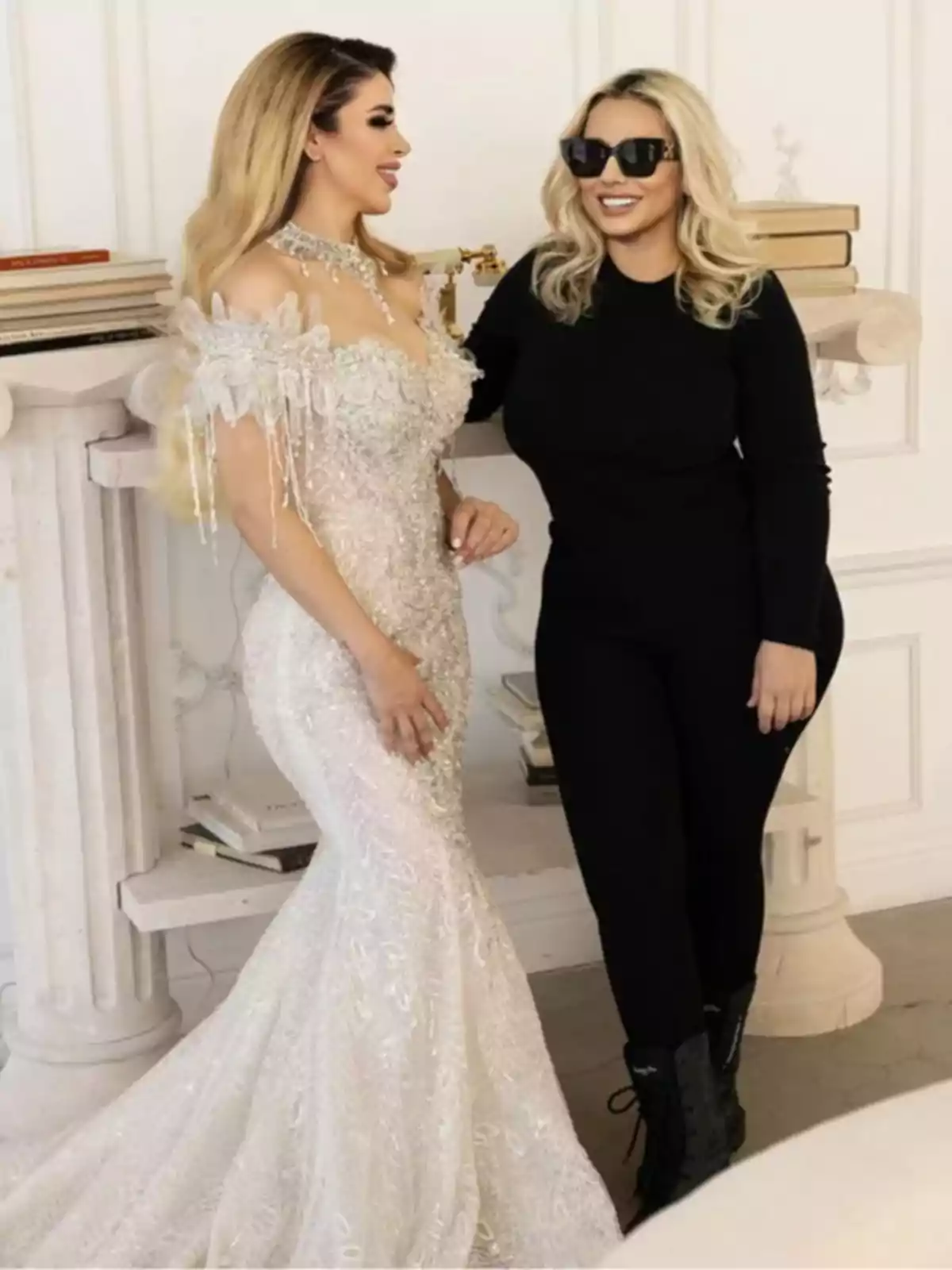 Dos mujeres posan juntas, una con un vestido de novia blanco y la otra con un atuendo negro y gafas de sol.