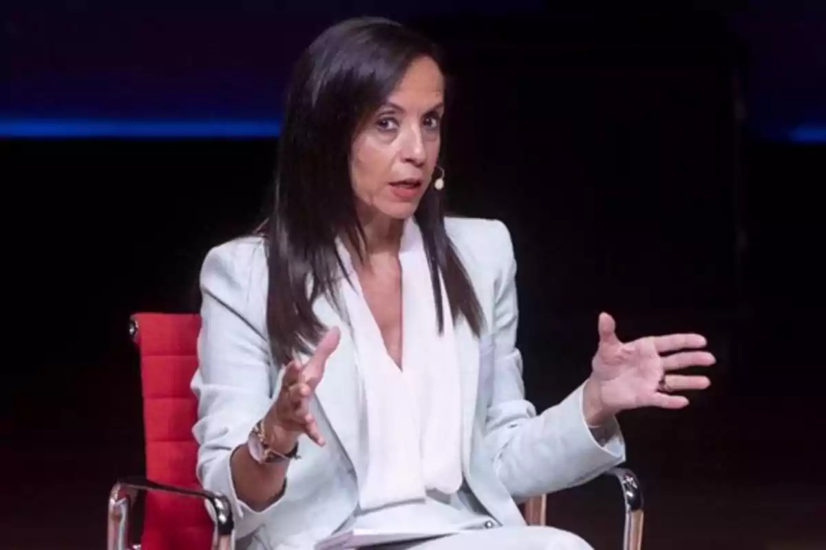 Una mujer con cabello oscuro y largo está sentada en una silla roja, gesticulando mientras habla en un escenario con fondo oscuro. Una mujer con cabello oscuro y largo está sentada en una silla roja, gesticulando mientras habla en un escenario con fondo oscuro.