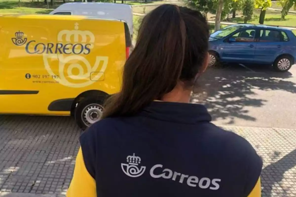 Trabajadora de Correos de espaldas frente a una furgoneta amarilla de la empresa en la calle Trabajadora de Correos de espaldas frente a una furgoneta amarilla de la empresa en la calle