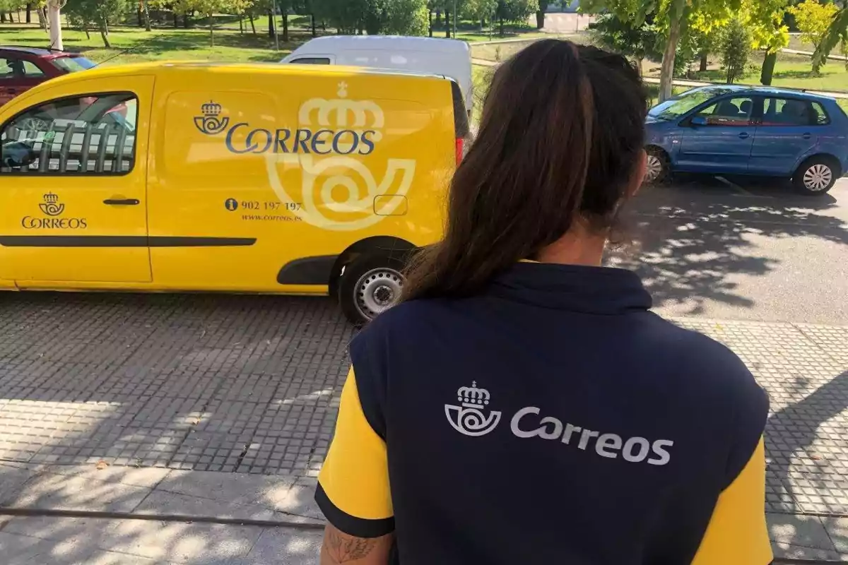 Una persona de espaldas con uniforme de Correos observa una furgoneta amarilla de la misma empresa estacionada en la calle.