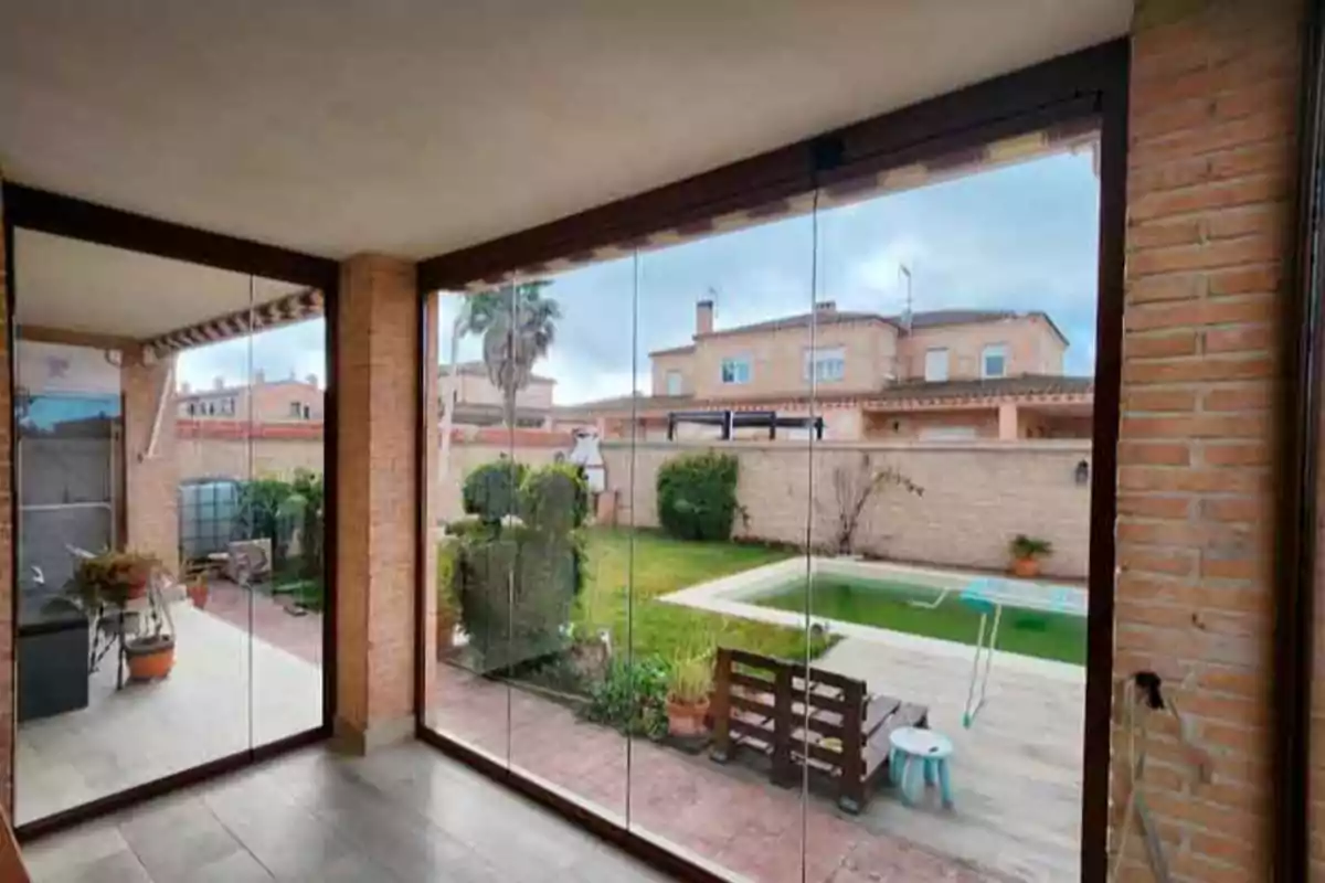 Vista desde un porche acristalado hacia un jardín con piscina y casas al fondo.