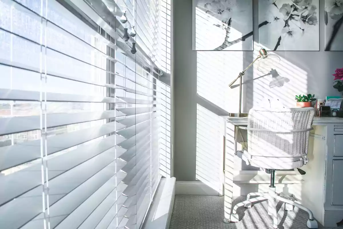 Oficina en casa iluminada por la luz del sol que entra a través de persianas blancas con un escritorio blanco una silla de mimbre y una lámpara dorada