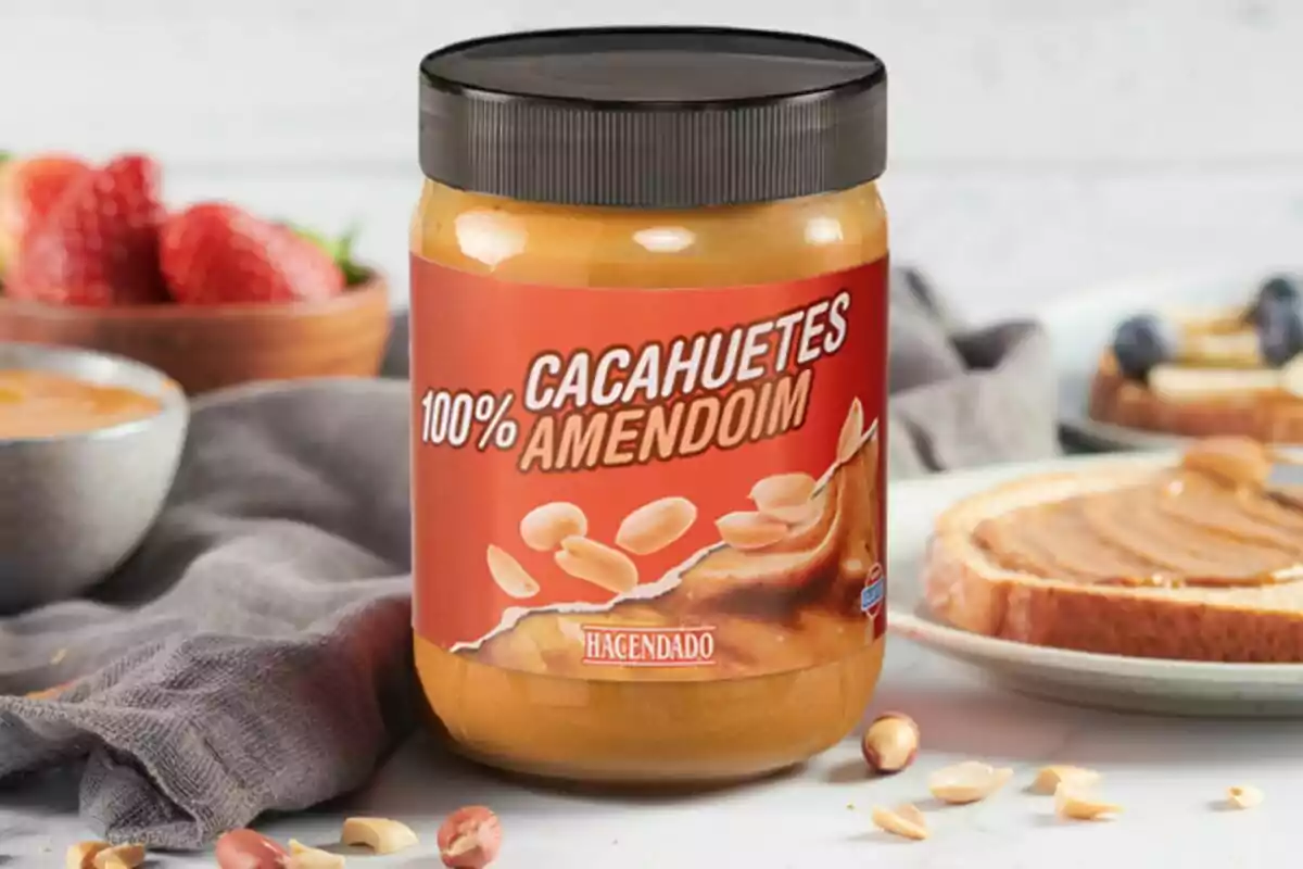 Un frasco de crema de cacahuate de Mercadona sobre una mesa con fresas y pan untado al fondo. Un frasco de crema de cacahuate de Mercadona sobre una mesa con fresas y pan untado al fondo.