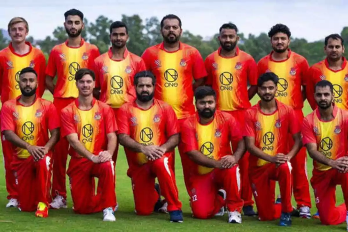 Un equipo de cricket posando para una foto grupal, todos llevan uniformes rojos y amarillos con un logotipo en el centro.