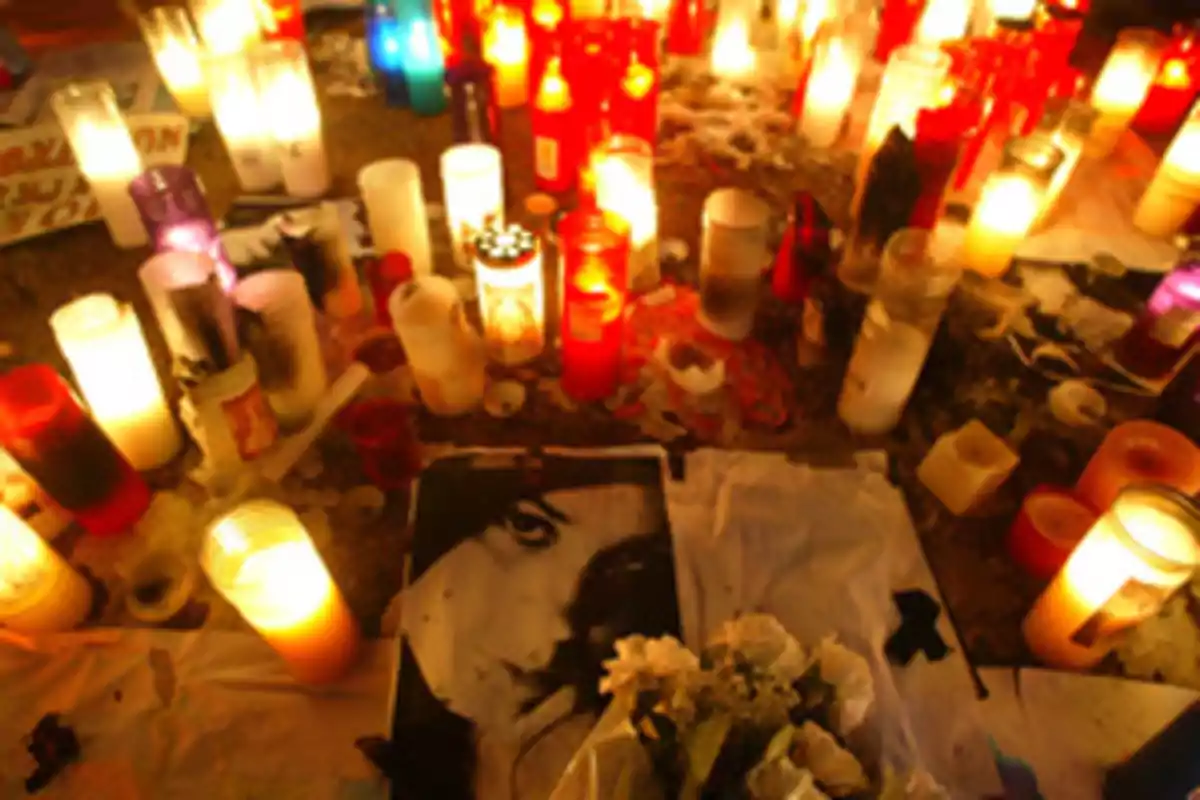 Un altar con velas encendidas de diferentes colores y tamaños rodea una fotografía en blanco y negro junto a un ramo de flores blancas.