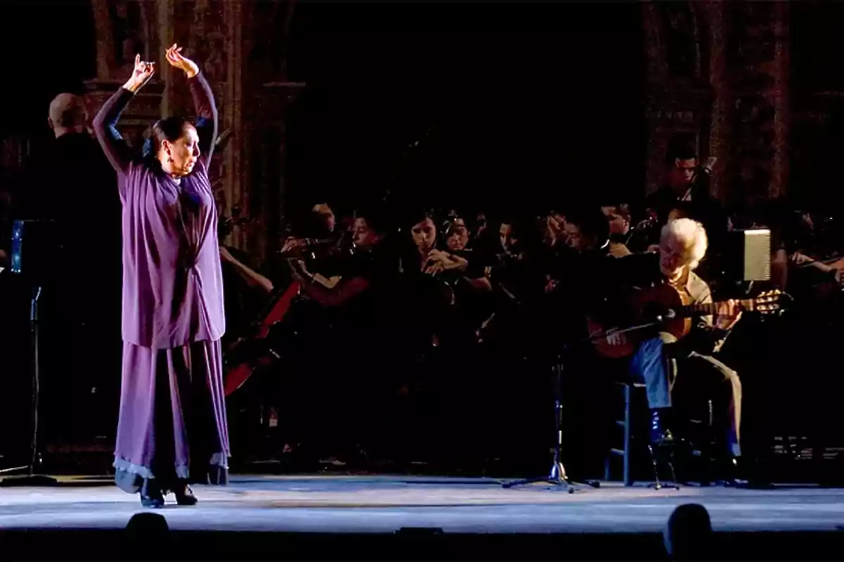 Una bailarina de flamenco con un vestido morado levanta los brazos mientras un guitarrista toca en el escenario, con una orquesta al fondo.