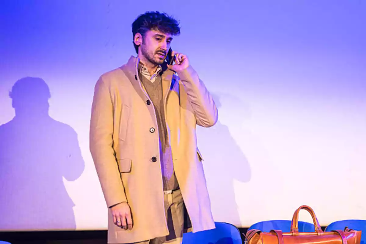 Un hombre con abrigo beige habla por teléfono en un escenario iluminado de azul, con una bolsa de cuero a su lado. Un hombre con abrigo beige habla por teléfono en un escenario iluminado de azul, con una bolsa de cuero a su lado.