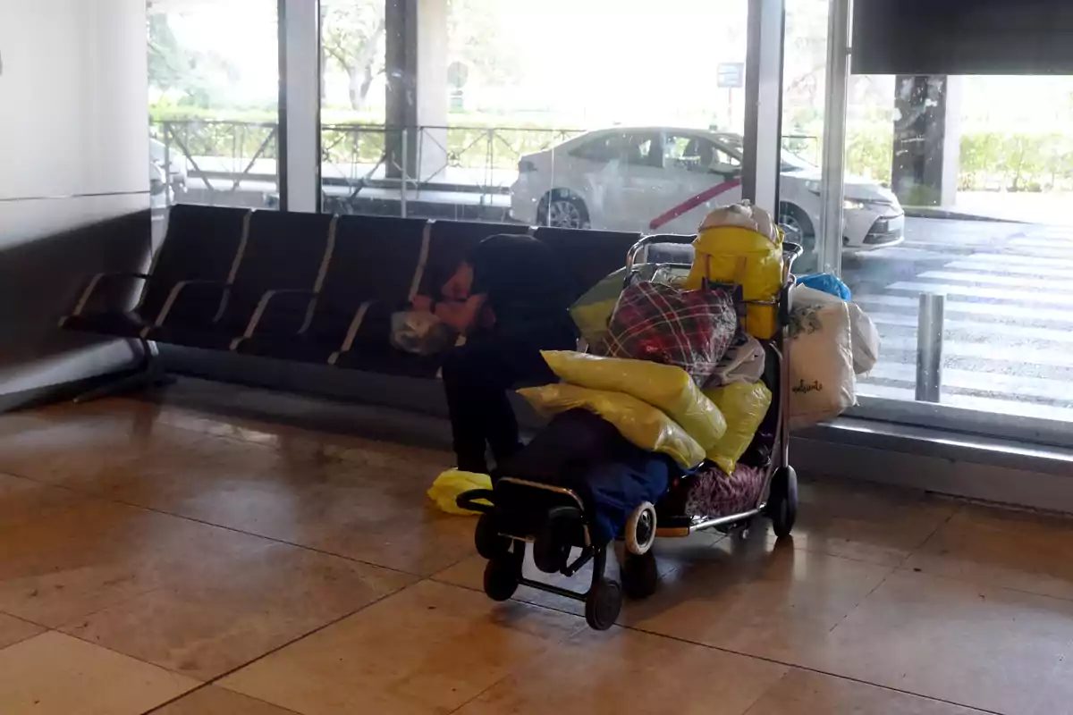 Una persona duerme en una banca de una estación o terminal, con un carrito lleno de pertenencias a su lado.