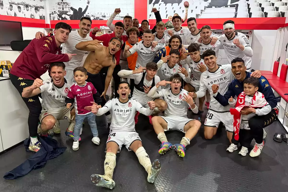 Un grupo de jugadores de fútbol celebra con entusiasmo en un vestuario después de un partido, algunos están de pie y otros sentados en el suelo, todos lucen uniformes blancos con el logo de Aspire Academy, hay dos niños pequeños entre ellos, uno de los cuales sostiene una bufanda roja.