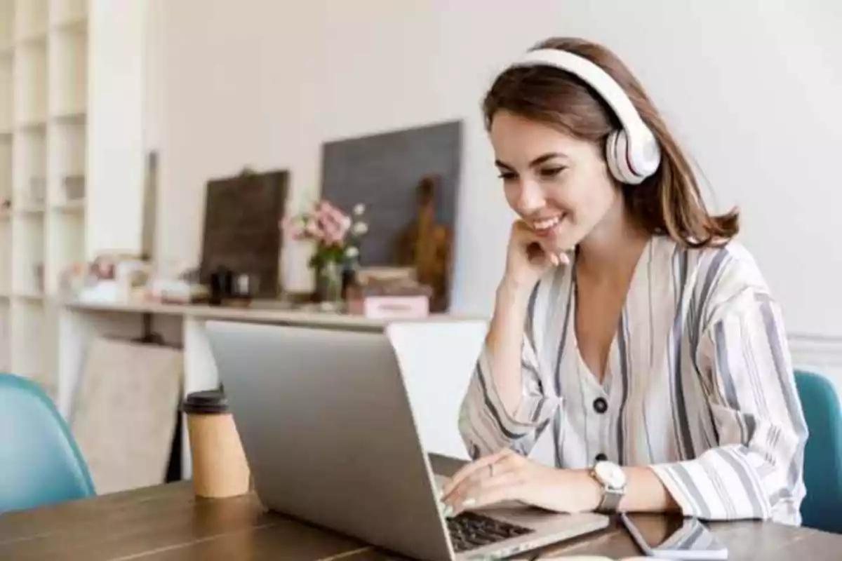 Mujer con auriculares trabajando en una computadora portátil en una mesa con una taza de café desechable.