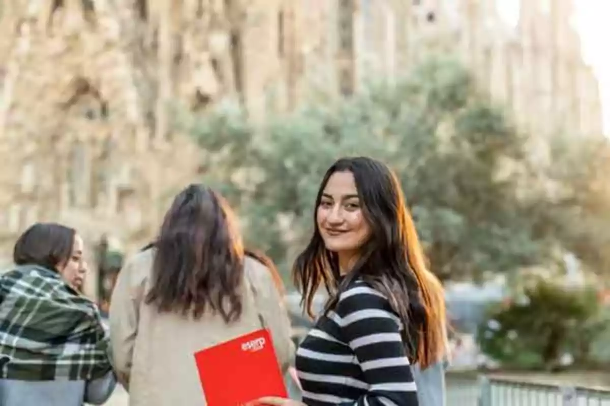 Una mujer sonriente con un suéter de rayas sostiene un cuaderno rojo mientras está de pie al aire libre con un edificio histórico de fondo.