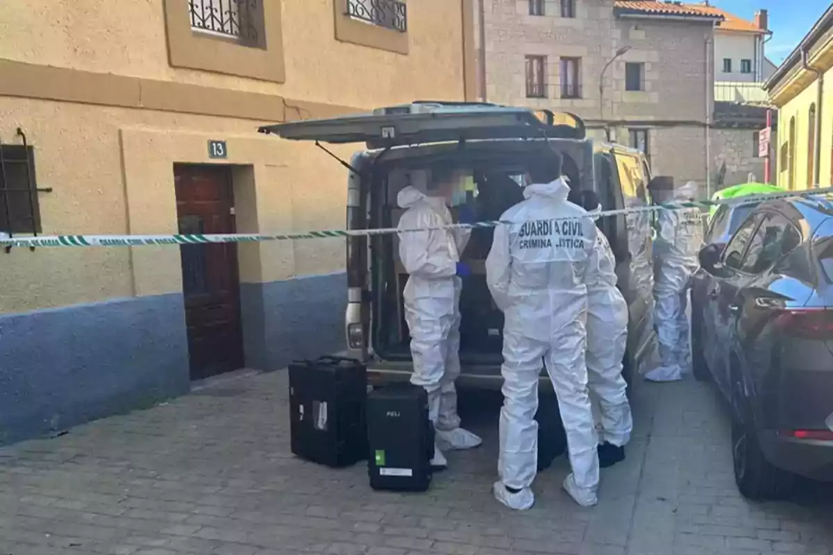 Un grupo de personas con trajes de protección de la Guardia Civil realiza una investigación en una calle acordonada frente a un edificio.