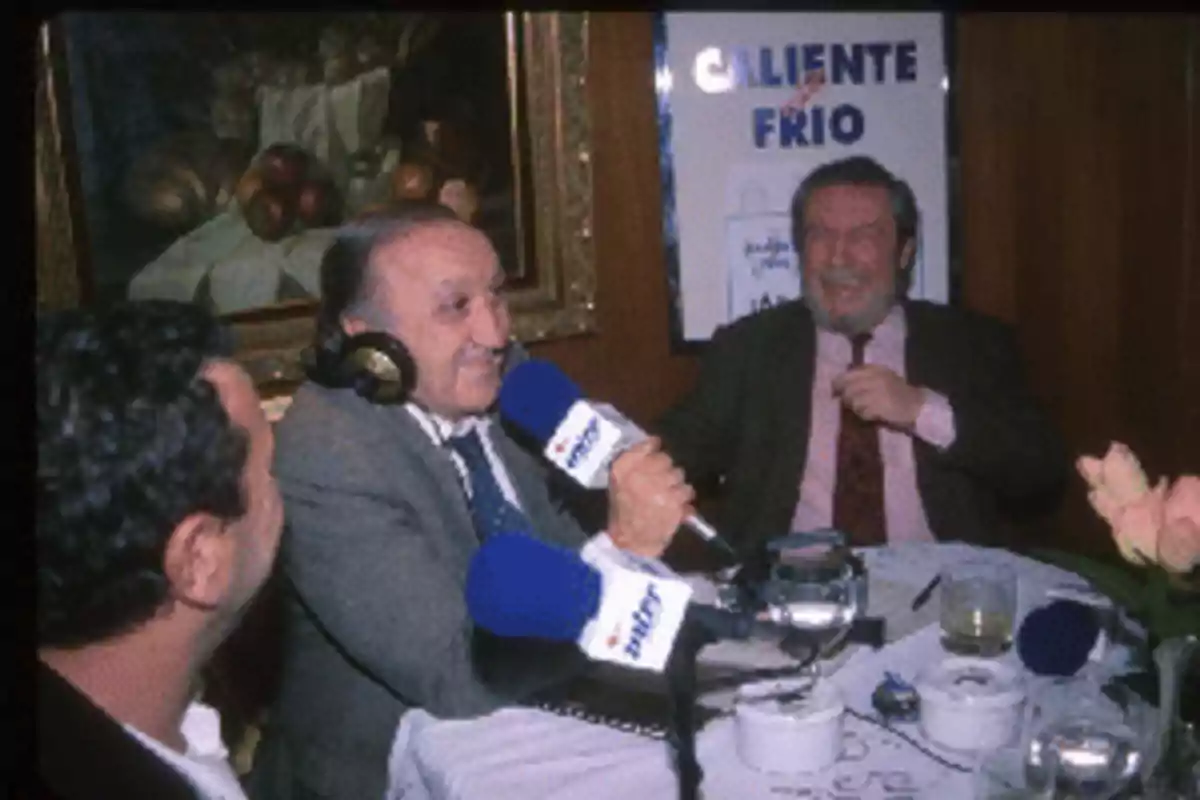 Tres hombres en una mesa de restaurante participan en una entrevista de radio, con micrófonos y auriculares, mientras sonríen y conversan animadamente.