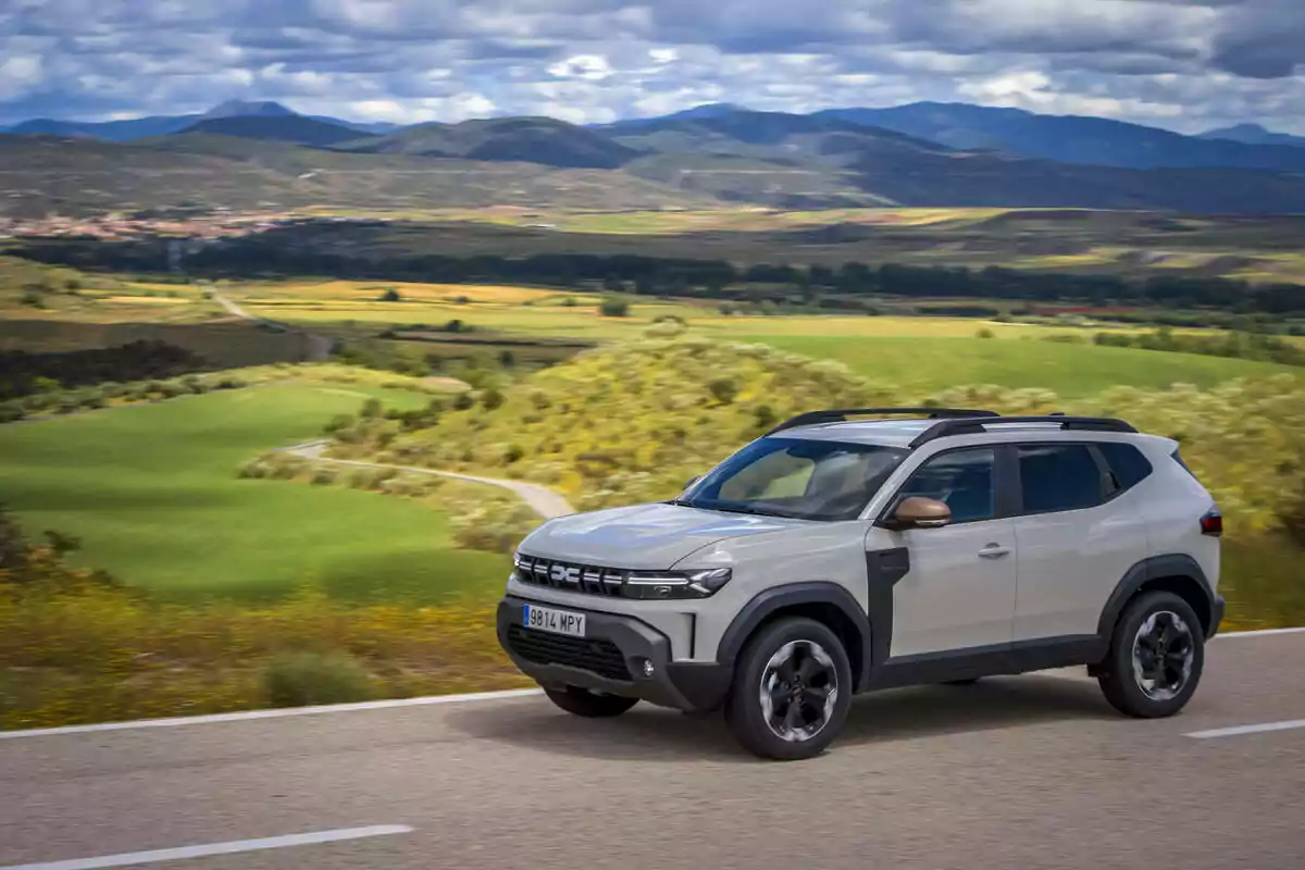 Un Dacia Duster Extreme Hybrid 140 blanco circula por una carretera en medio de un paisaje rural con colinas verdes y montañas al fondo bajo un cielo parcialmente nublado Un Dacia Duster Extreme Hybrid 140 blanco circula por una carretera en medio de un paisaje rural con colinas verdes y montañas al fondo bajo un cielo parcialmente nublado