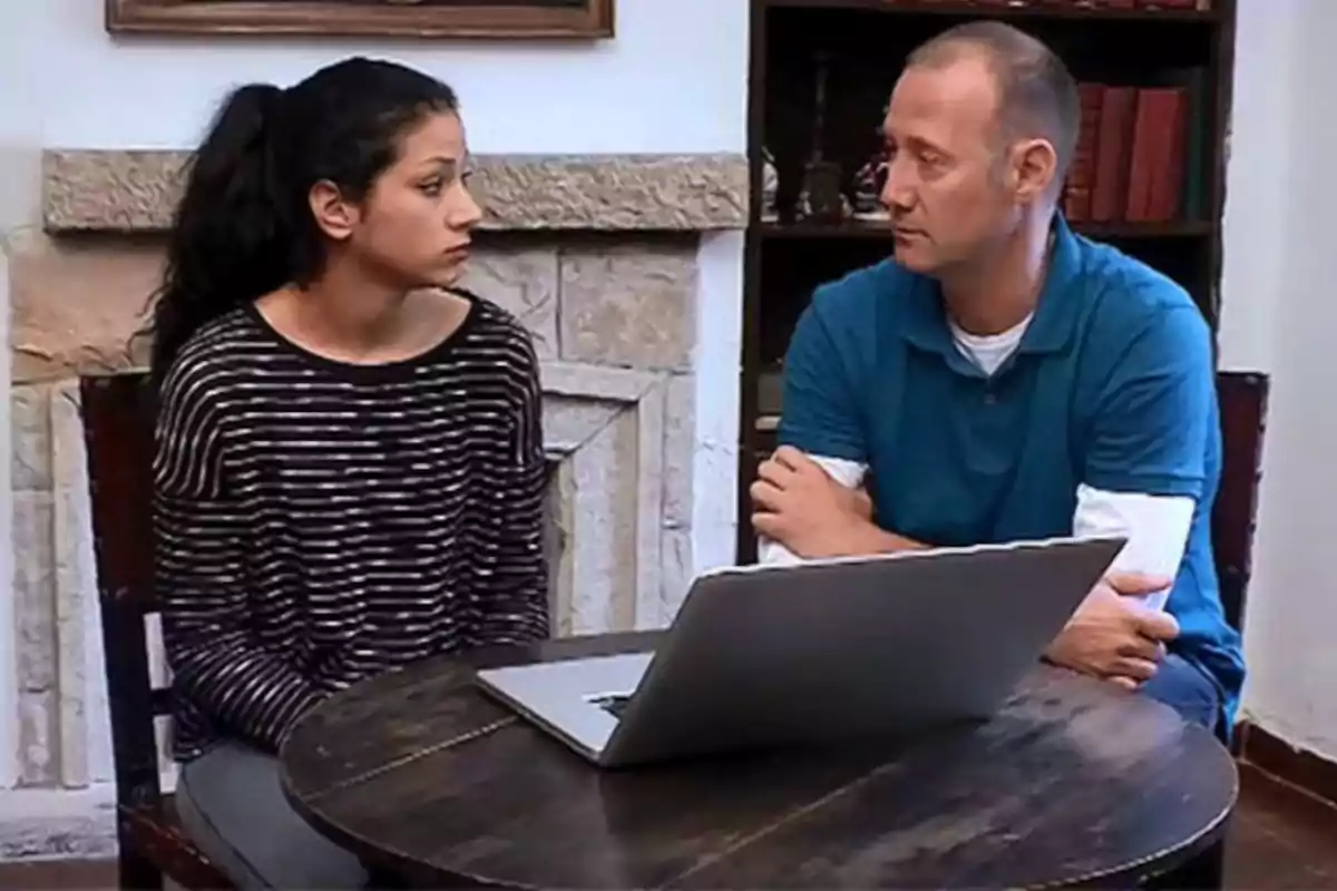 Una mujer y un hombre sentados en una mesa redonda con una computadora portátil, mirándose el uno al otro en una conversación seria.