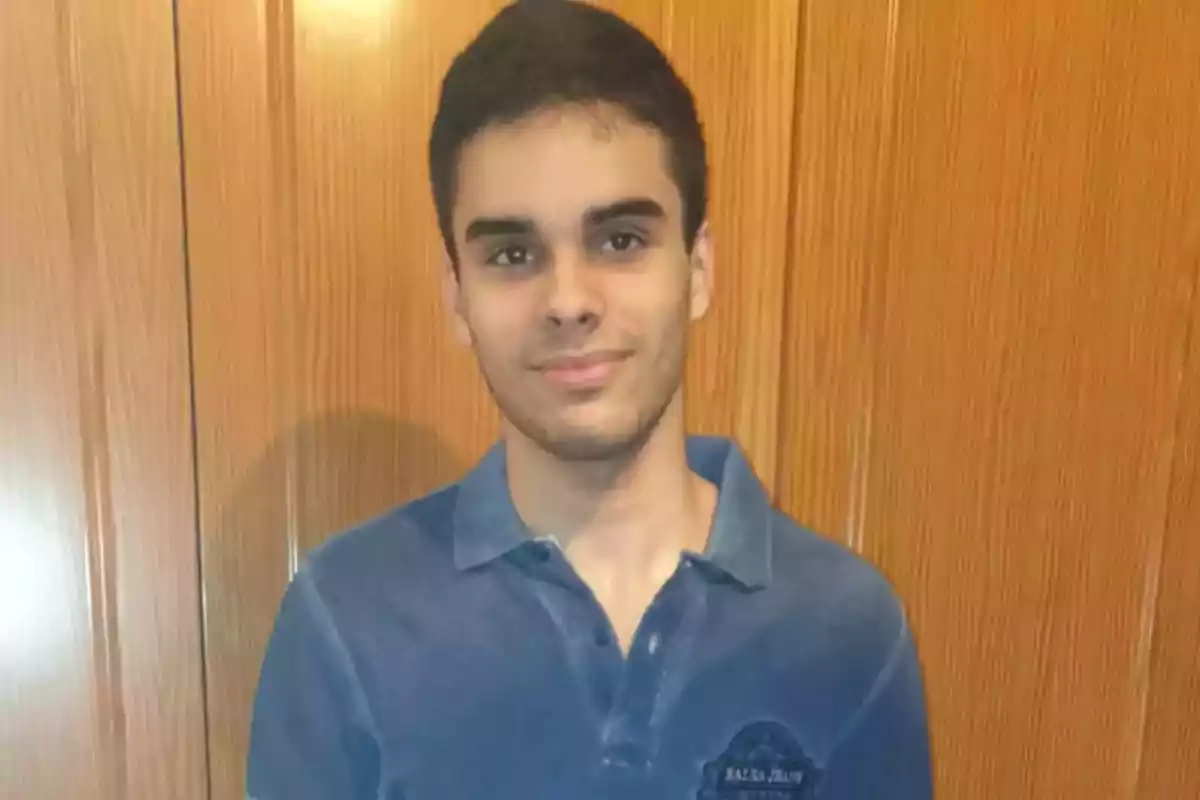 Un joven con una camisa azul de pie frente a una puerta de madera.