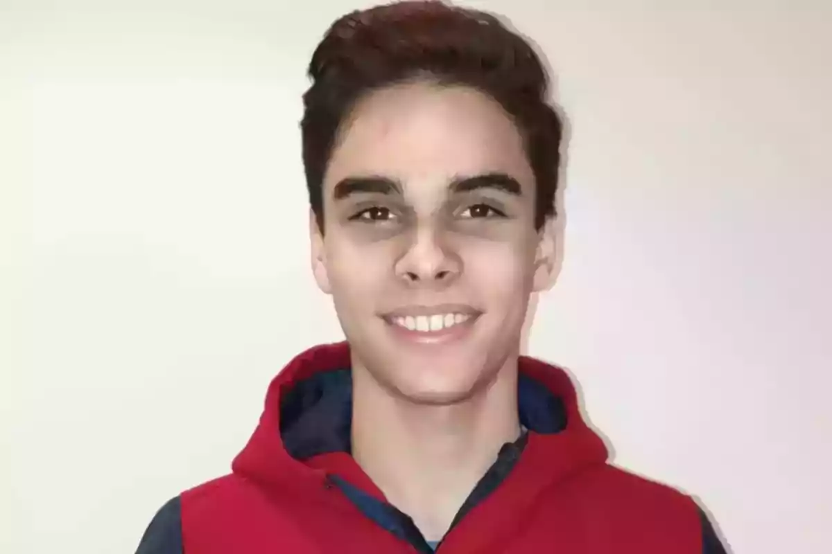 Un joven con cabello oscuro y corto sonríe a la cámara mientras lleva una sudadera roja con capucha.