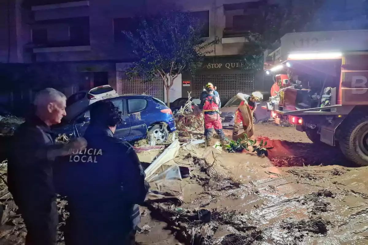 Un grupo de bomberos y policías locales trabaja en una calle llena de barro y escombros tras una inundación, con un coche dañado y un camión de bomberos iluminando la escena.