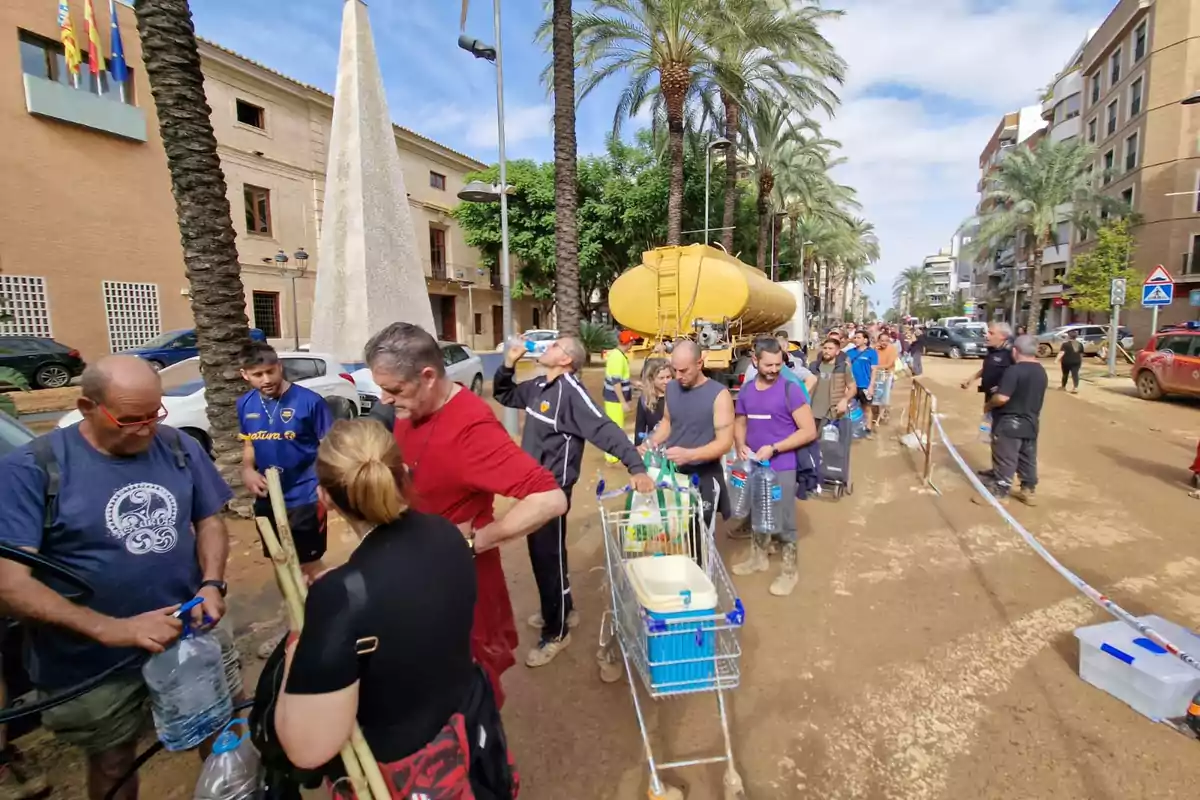 Personas haciendo fila en una calle para recoger agua de un camión cisterna amarillo, rodeados de palmeras y edificios.