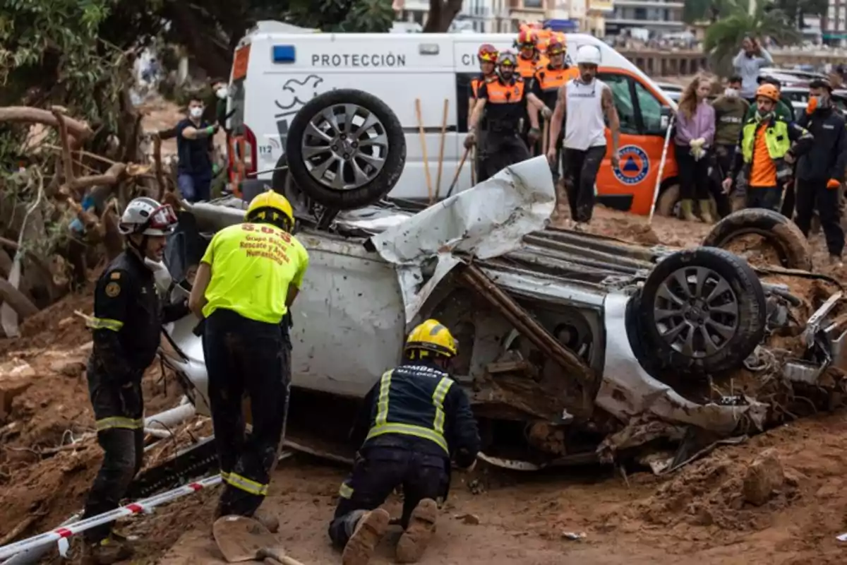 Rescatistas trabajan alrededor de un coche volcado en una zona afectada por un desastre natural, con un vehículo de protección civil y más personal de emergencia al fondo.