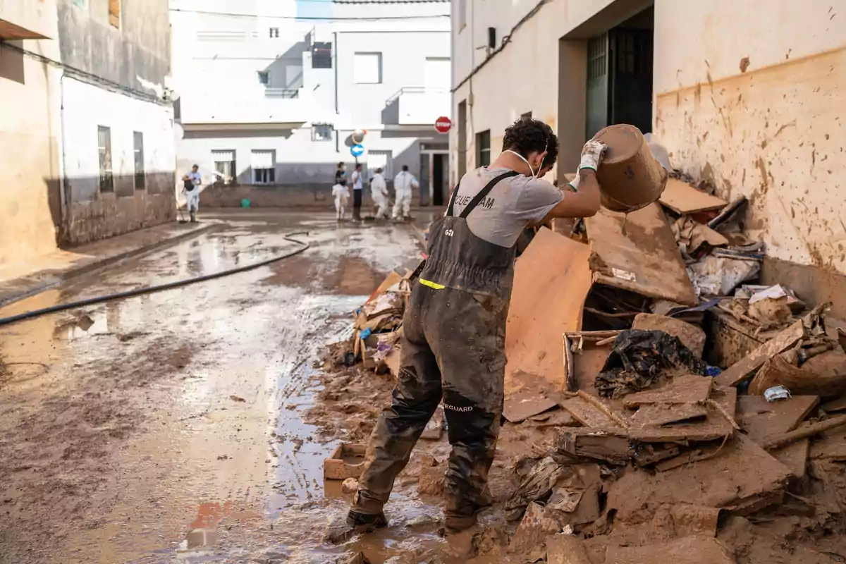 Un hombre con botas y guantes limpia escombros y barro en una calle después de una inundación.