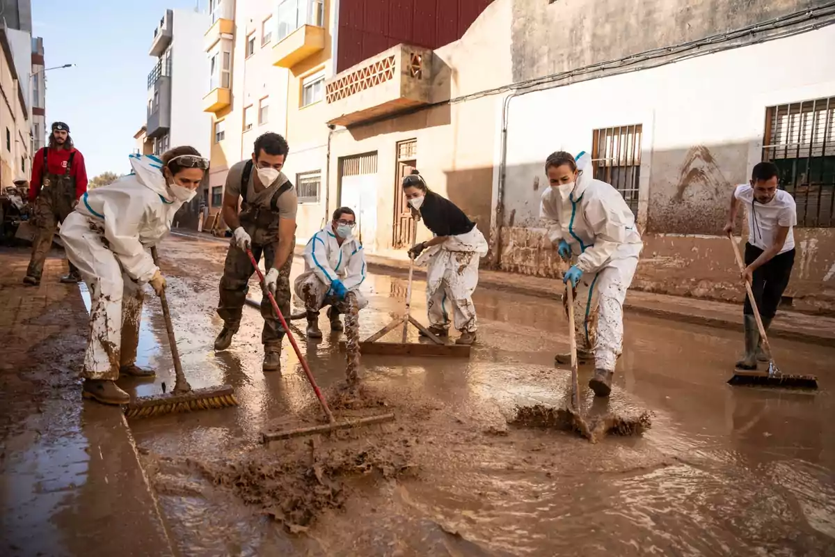 Un grupo de personas con equipo de protección y mascarillas limpian una calle llena de barro después de una inundación.