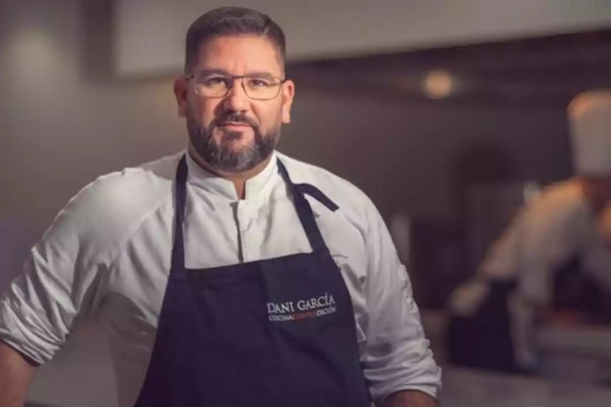 Un chef con gafas y barba, vestido con uniforme de cocina y delantal, en un entorno de cocina profesional.