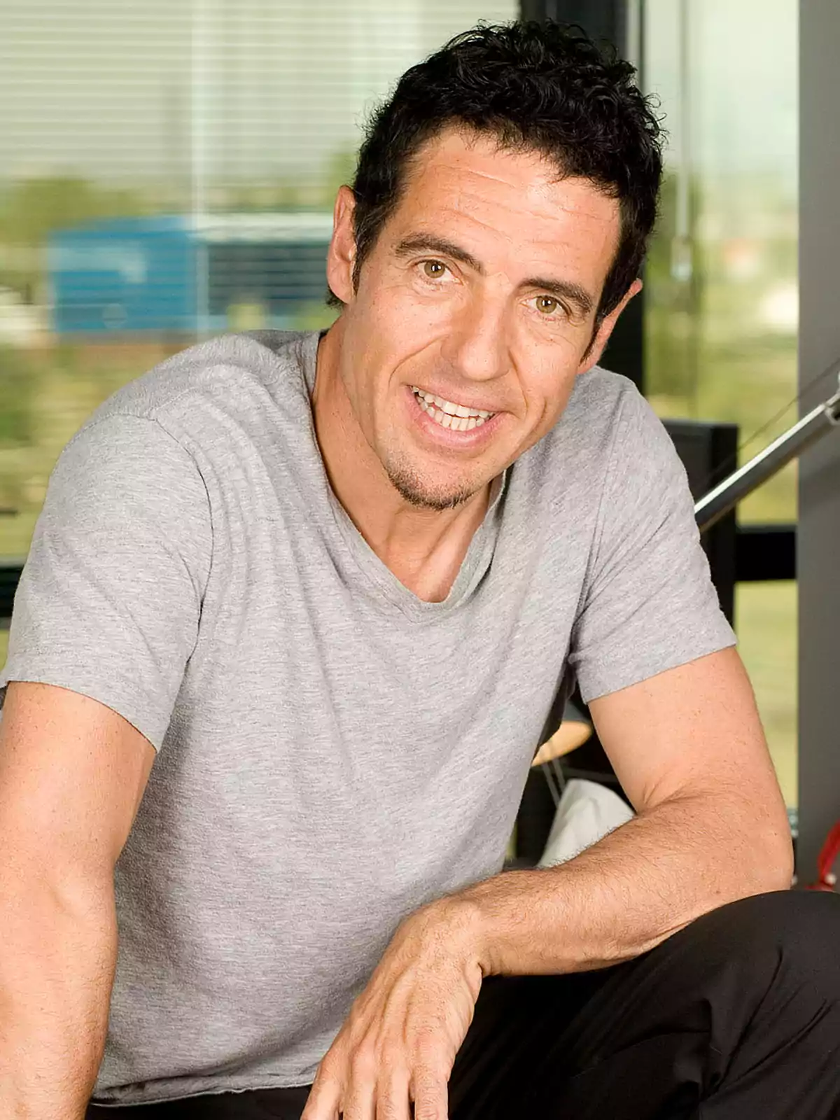 Hombre de cabello corto y oscuro con camiseta gris sonriendo y sentado en un ambiente interior moderno Hombre de cabello corto y oscuro con camiseta gris sonriendo y sentado en un ambiente interior moderno