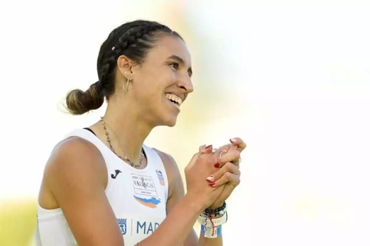 Una atleta sonriente con el cabello trenzado y las manos juntas, vistiendo una camiseta deportiva blanca. Una atleta sonriente con el cabello trenzado y las manos juntas, vistiendo una camiseta deportiva blanca.