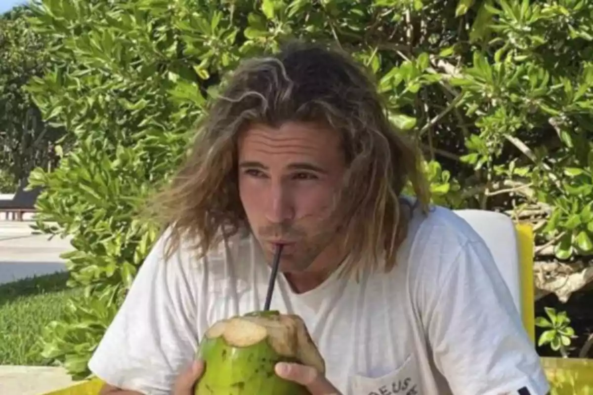 Hombre con cabello largo bebiendo de un coco con pajilla en un entorno tropical.