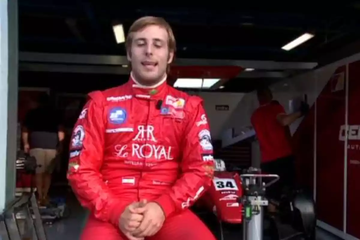 Piloto de automovilismo con traje rojo sentado frente a un garaje de boxes con un monoplaza de carreras al fondo Piloto de automovilismo con traje rojo sentado frente a un garaje de boxes con un monoplaza de carreras al fondo