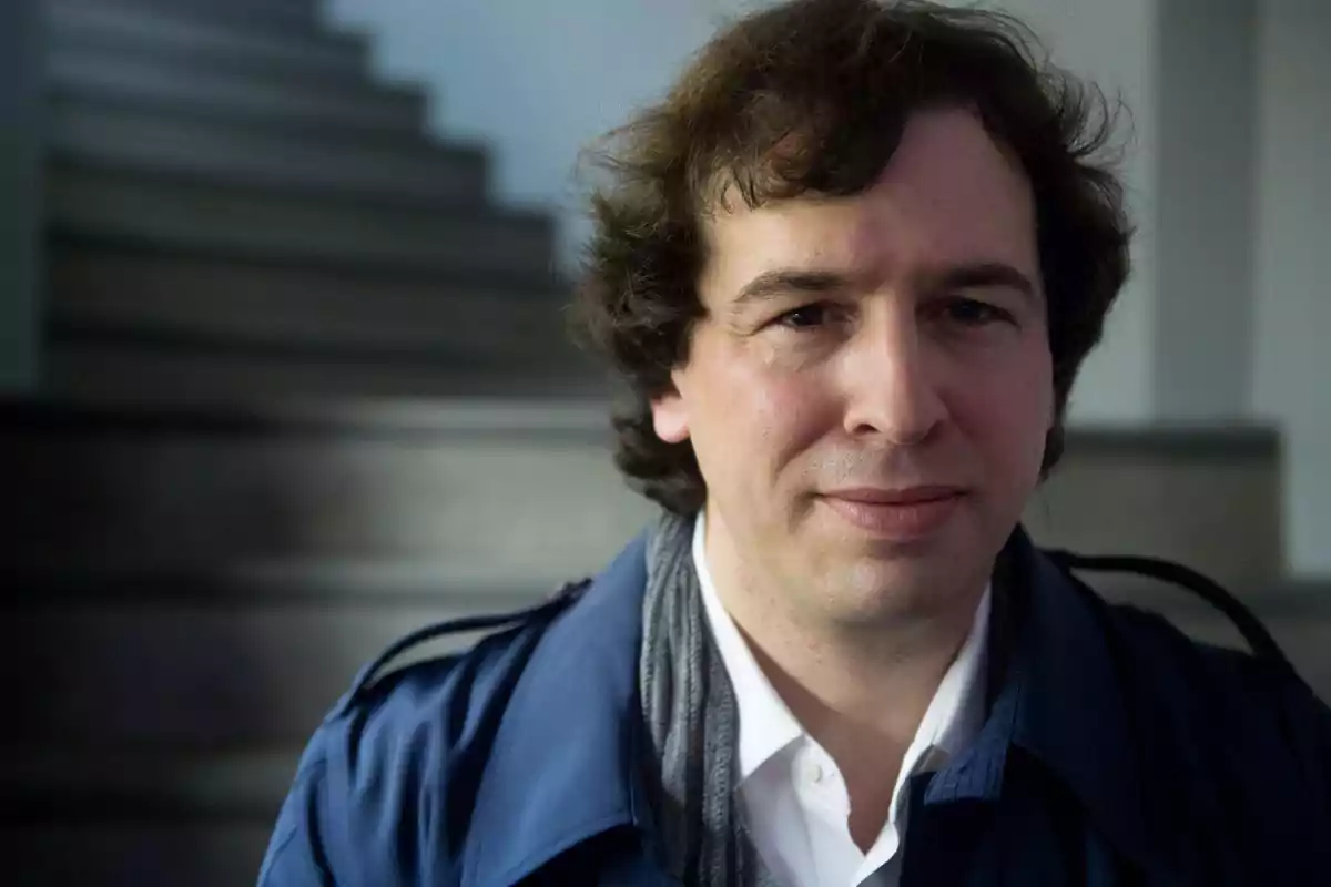 Hombre con cabello rizado y chaqueta azul sonriendo frente a una escalera borrosa.