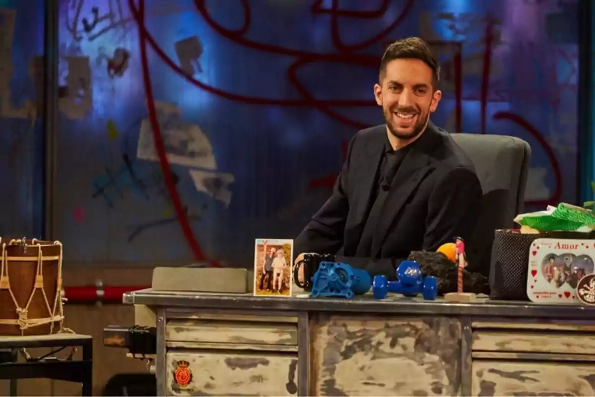Un hombre sonriente con barba y traje oscuro está sentado detrás de un escritorio desordenado con varios objetos, incluyendo una foto, juguetes y una caja con la palabra 