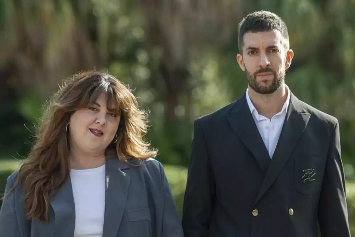 Lalachus y David Broncano posan al aire libre con trajes formales y un fondo de vegetación.