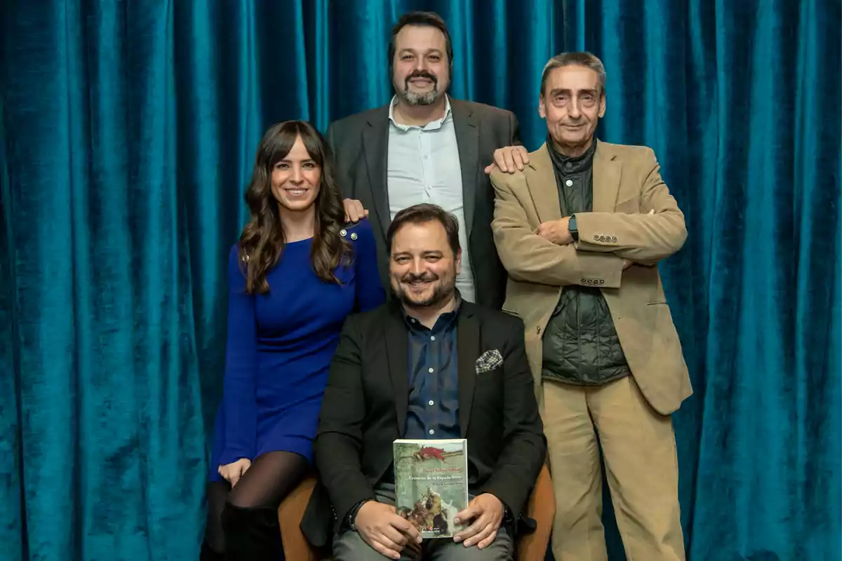 Un grupo de cuatro personas posando frente a una cortina azul, una mujer y tres hombres, uno de ellos sostiene un libro.