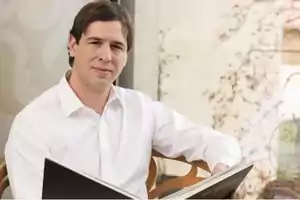 Hombre con camisa blanca sentado y sosteniendo un libro en un entorno interior con una ventana al fondo.