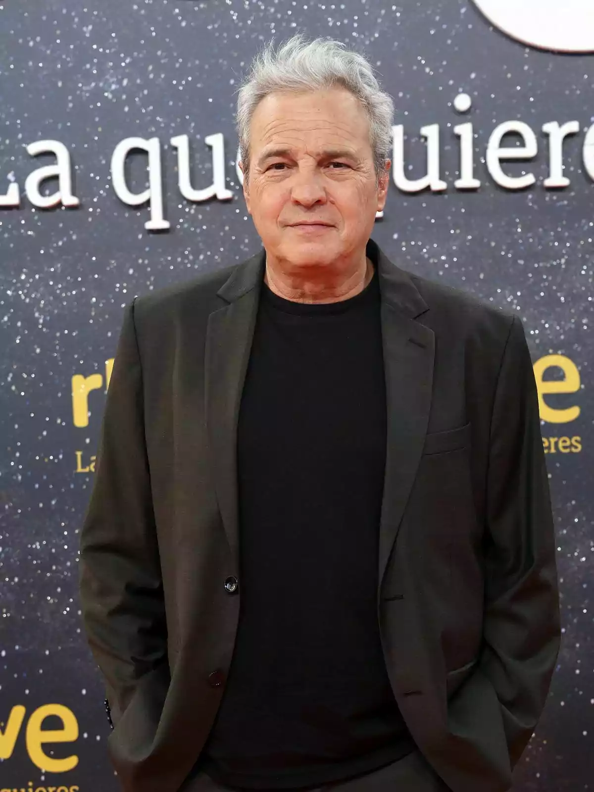 David Summers de expresión seria, vestido con un traje oscuro y camiseta negra, posa frente a un fondo estrellado con letras blancas y amarillas.