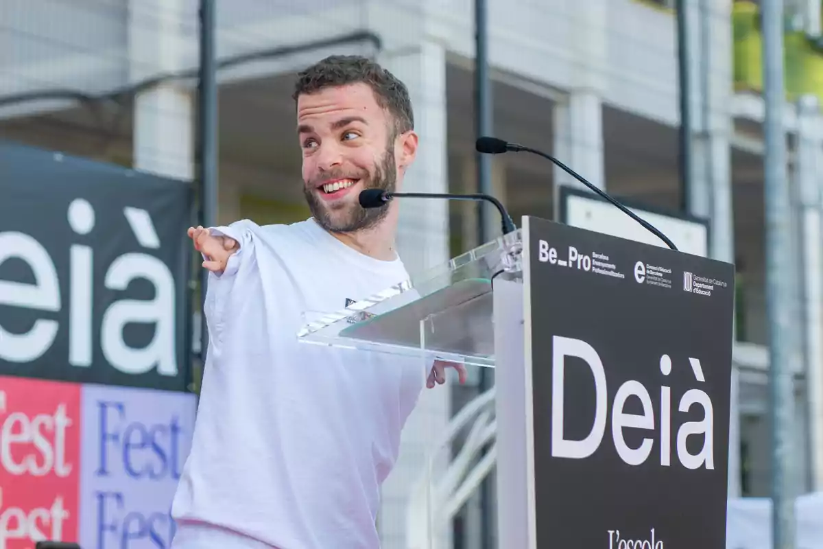 Un hombre sonriente con barba corta y camiseta blanca habla en un podio con micrófonos durante un evento al aire libre.