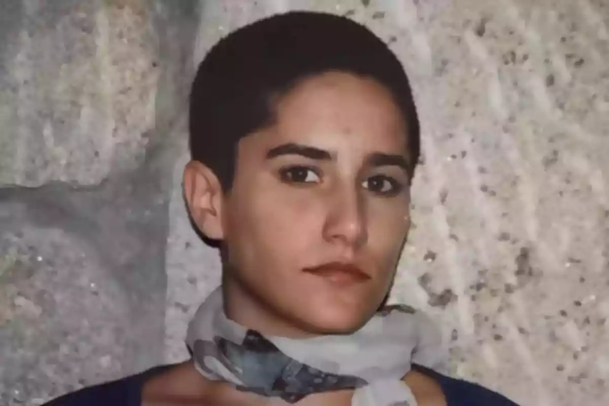Persona de cabello corto y oscuro con bufanda clara posando frente a una pared de piedra Persona de cabello corto y oscuro con bufanda clara posando frente a una pared de piedra