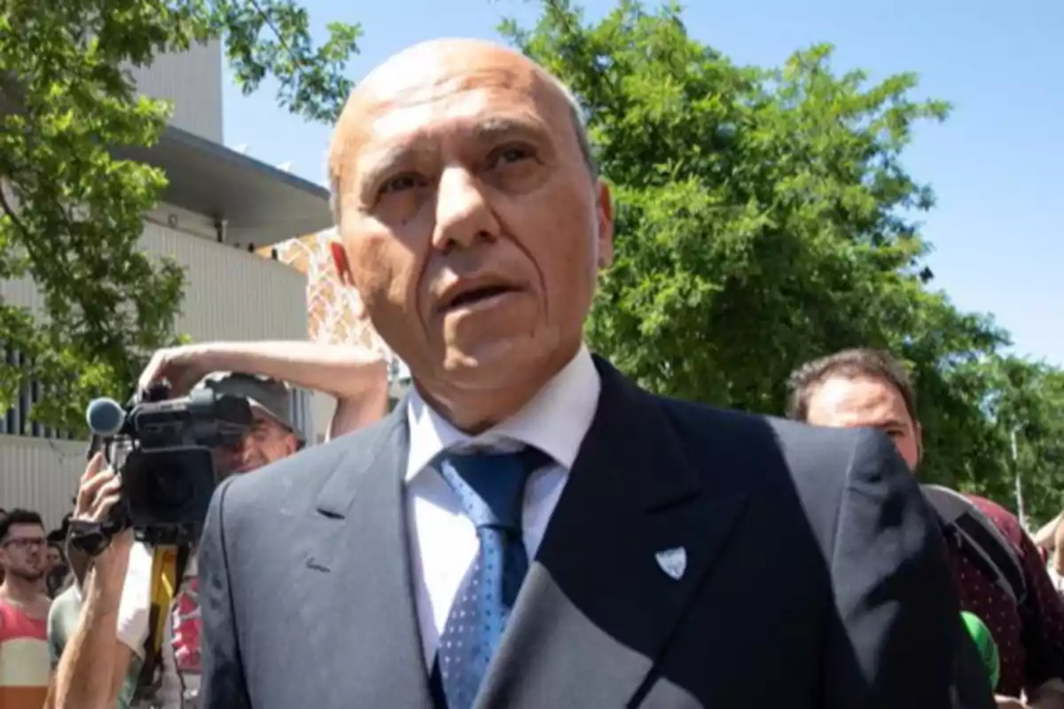 Un hombre calvo con traje oscuro y corbata azul está al aire libre rodeado de personas y cámaras, con árboles y un edificio de fondo. Un hombre calvo con traje oscuro y corbata azul está al aire libre rodeado de personas y cámaras, con árboles y un edificio de fondo.