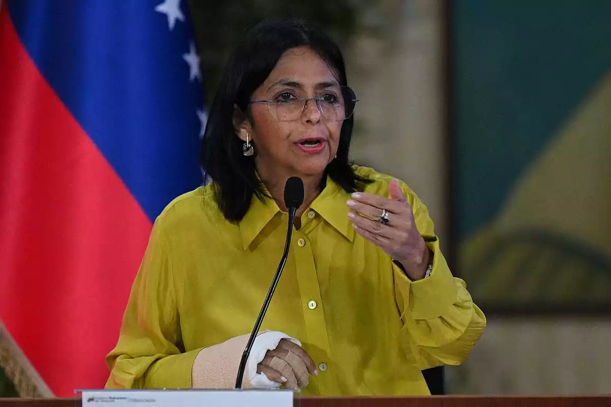 Una mujer con gafas y camisa amarilla habla frente a un micrófono con una bandera de fondo.