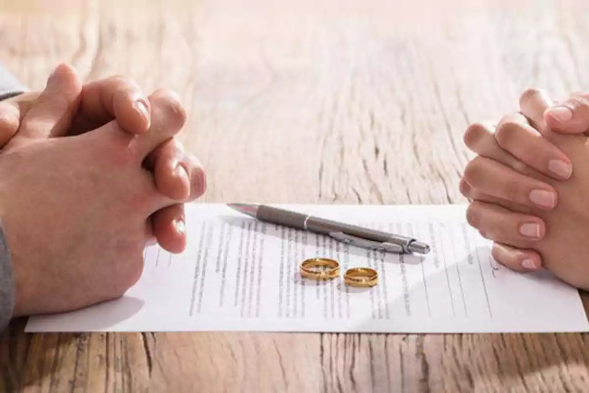Manos de una pareja frente a un documento con dos anillos y un bolígrafo sobre una mesa de madera.