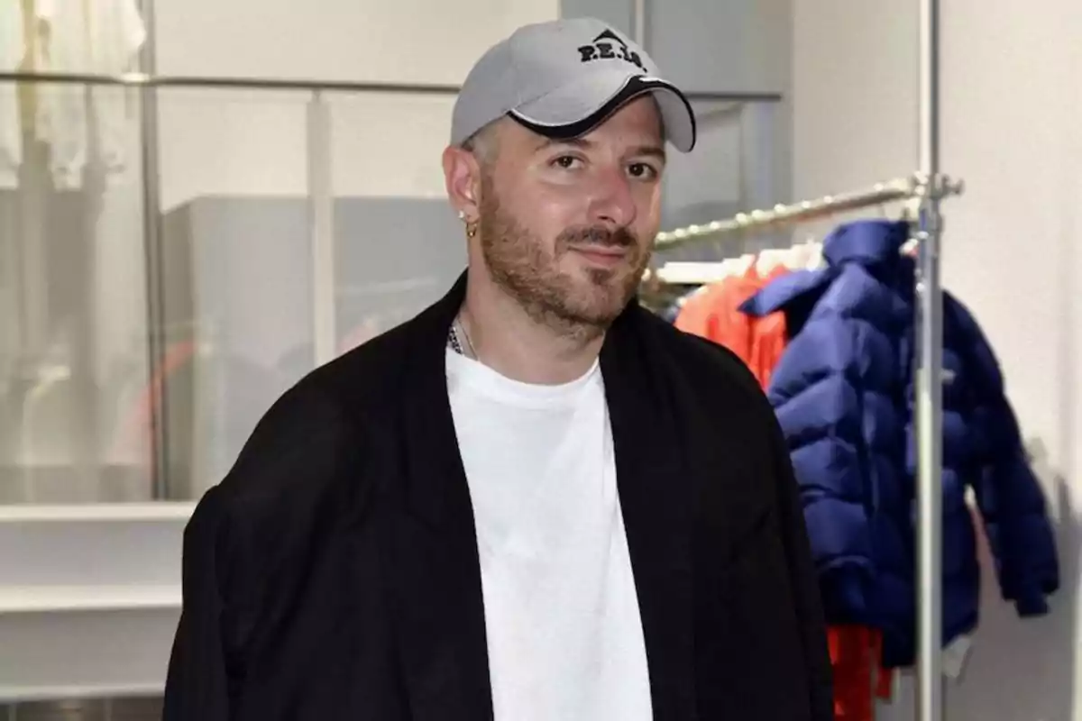 Un hombre con barba y gorra gris está de pie en una tienda de ropa. Lleva una camiseta blanca y una chaqueta negra. Al fondo, se pueden ver varias prendas colgadas, incluyendo chaquetas de colores brillantes.